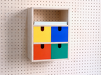 Veelkleurige opbergbox met 3 vakken aan pegboard: 4 lades in geel, blauw, rood en groen en een vak voor A4-papier
